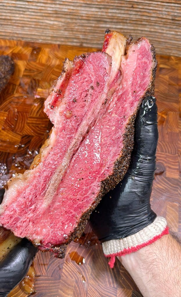 A gloved hand holds a thick, juicy slice of smoked beef brisket with a pink, tender interior and a dark, seasoned crust, displayed over a wooden cutting board.