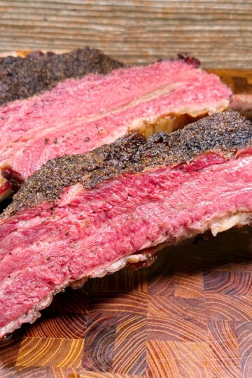 Two large, thick-cut smoked beef ribs with a dark, seasoned bark and a juicy pink interior rest on a wooden cutting board.