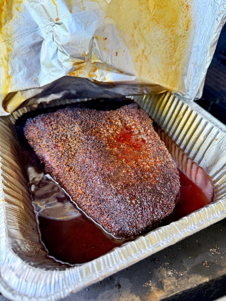 A cooked, seasoned brisket sits in a disposable aluminum tray filled with juices, partially covered by crumpled foil. The brisket has a dark, flavorful crust from the rub.