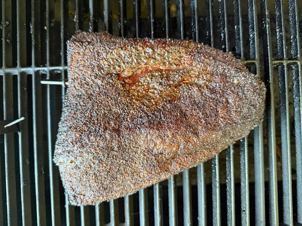 A large, seasoned beef brisket with a dark, crispy bark is being smoked on a grill, resting on the grates with visible grill bars beneath it.