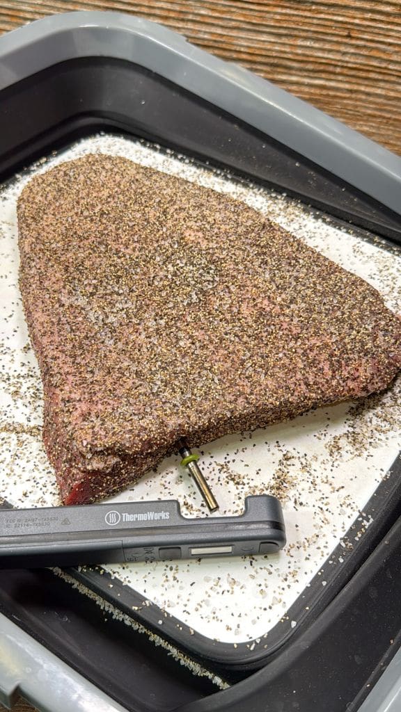 A seasoned brisket sits on parchment paper in a tray, with a digital meat thermometer inserted into the side to check its internal temperature.