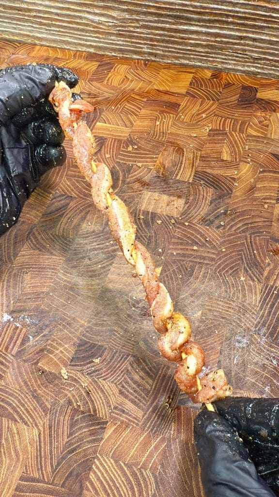 A person wearing black gloves holds a skewer with pieces of seasoned, twisted meat over a wooden cutting board.  These are bacon twists.