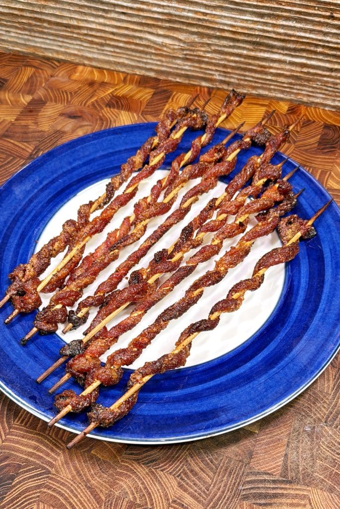 Several skewers of twisted, grilled meat are arranged on a blue plate with a white center, set on a wooden surface with a textured background.