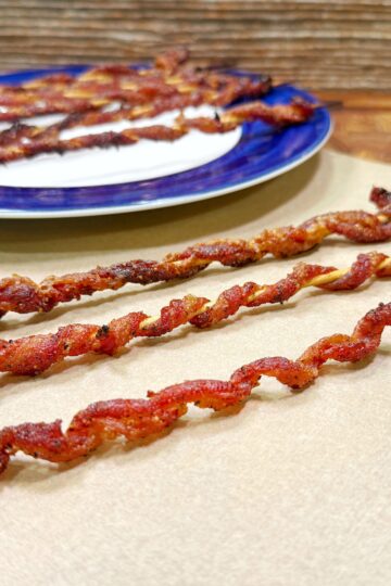 Four twisted strips of cooked bacon are skewered on sticks and laid out on parchment paper, with more bacon skewers on a blue-rimmed plate in the background. The surface and backdrop are wooden.