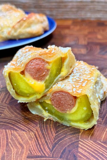 A close-up of two pastry-wrapped hot dogs with visible pickles and melted cheese inside, topped with sesame seeds, stacked on a wooden surface. A whole pastry is blurred in the background on a blue plate.