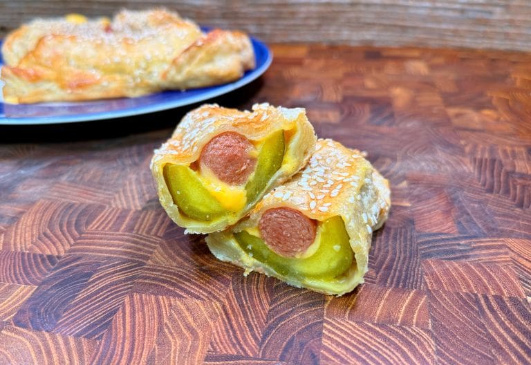 A close-up of two pastry-wrapped hot dogs with visible pickles and melted cheese inside, topped with sesame seeds, stacked on a wooden surface. A whole pastry is blurred in the background on a blue plate.