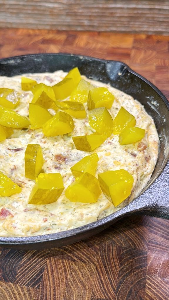 A skillet filled with a creamy dip, topped with chunks of bright yellow pickles, sits on a wooden surface.