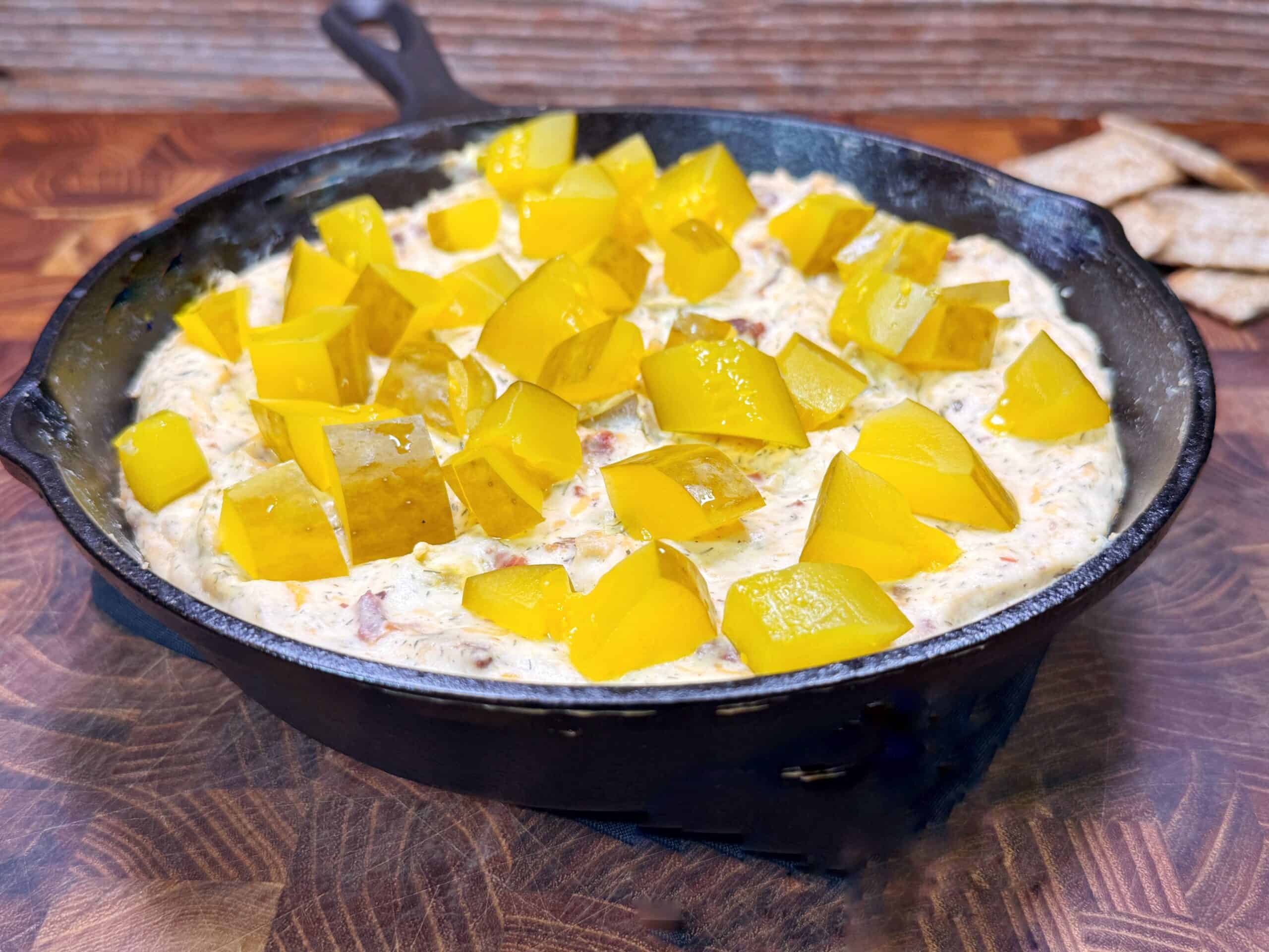 A cast iron skillet filled with creamy dip topped with diced pickles, set on a wooden surface with some crackers nearby.