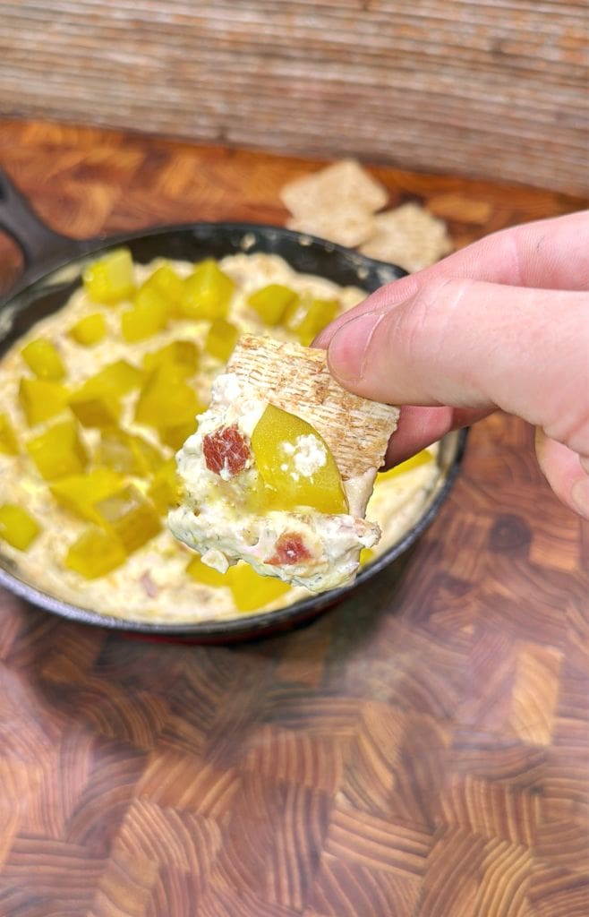 A hand holds a cracker topped with creamy dip, bacon bits, and diced pickles above a skillet filled with more dip and large pickle chunks, on a wooden surface.