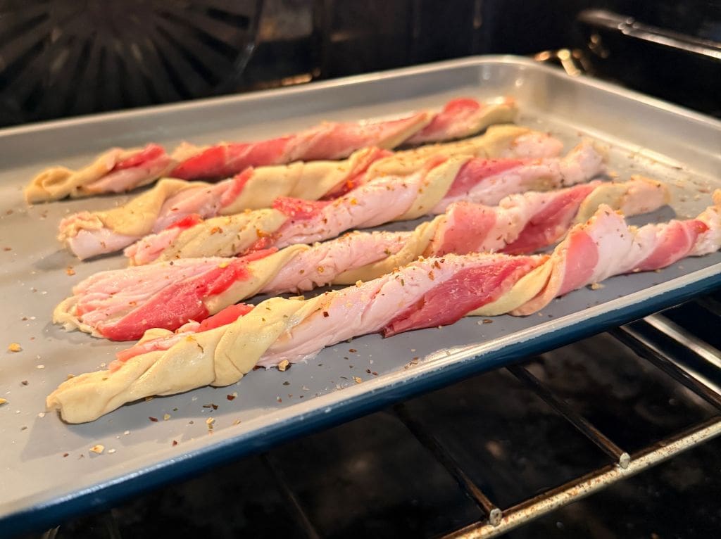 Raw bacon and dough twists arranged in rows on a baking sheet, sprinkled with seasoning, and placed inside an oven for baking.
