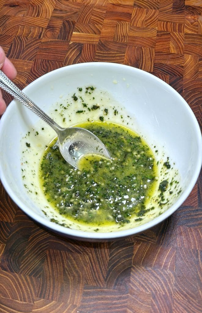 A hand holds a spoon in a white bowl containing a green and yellow herb mixture, likely a sauce or marinade, on a wooden surface.