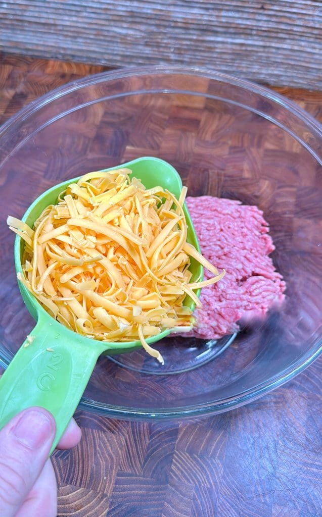 A hand holds a green measuring cup filled with shredded cheddar cheese above a glass bowl containing raw ground beef, on a wooden surface.