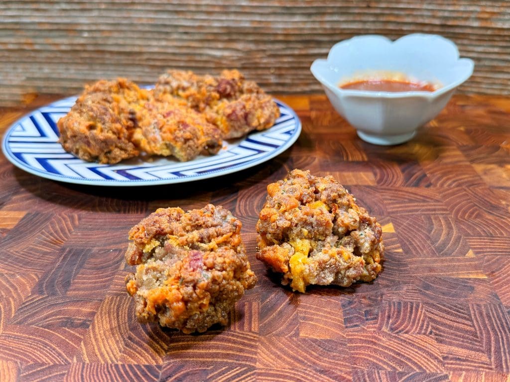 Two sausage balls sit on a wooden surface, with more sausage balls on a patterned plate and a small bowl of dipping sauce in the background.