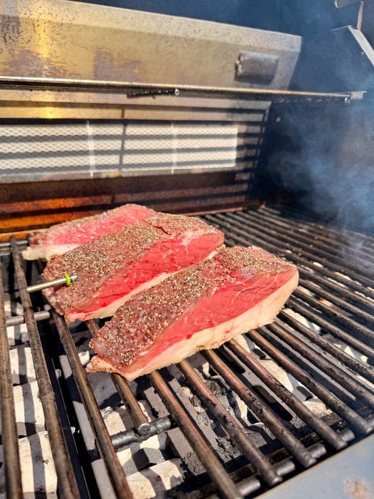 Three thick, seasoned steaks are cooking on a grill with visible grill marks and smoke rising, suggesting they are being cooked outdoors on a barbecue.