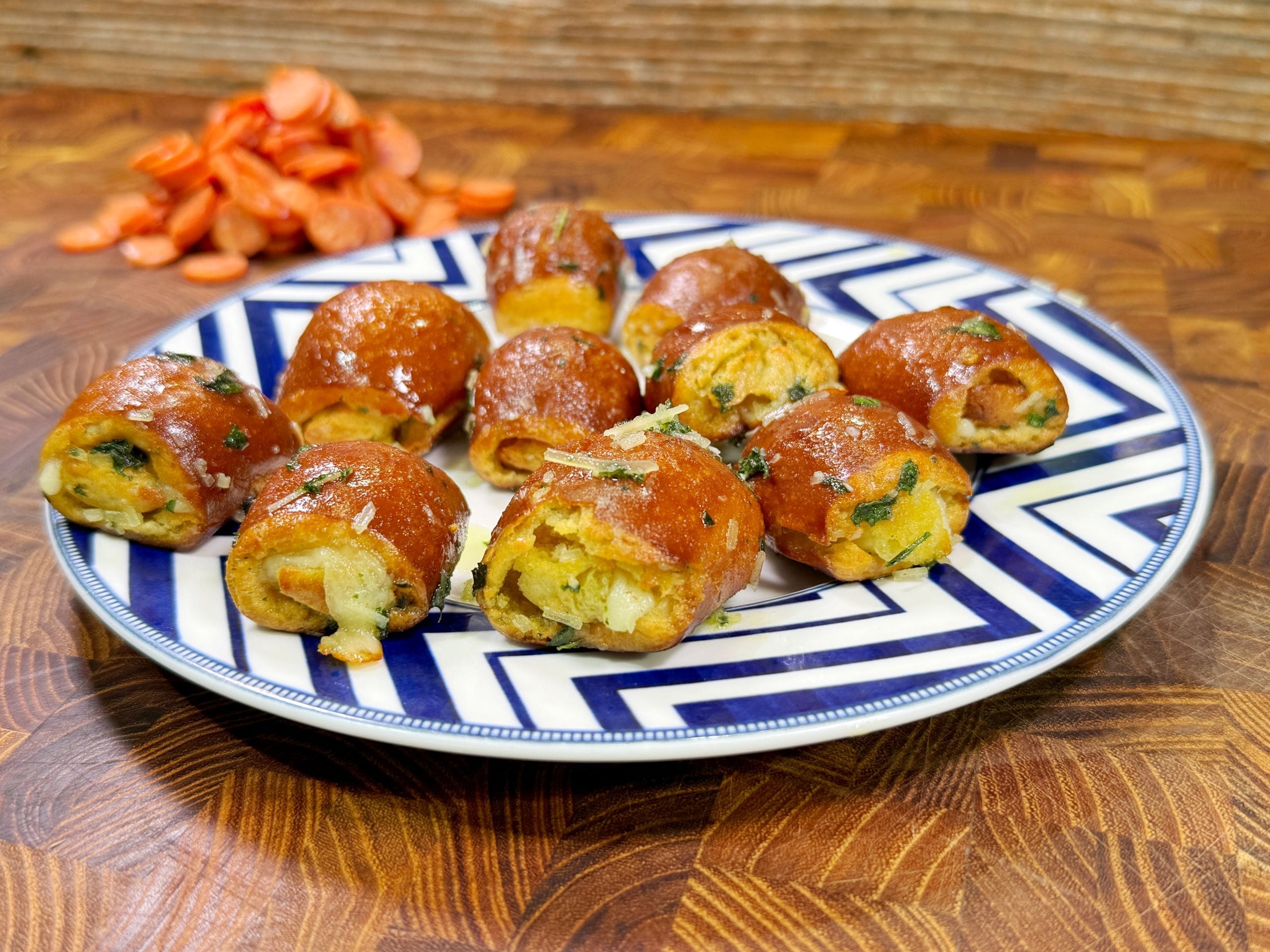 A plate with round, golden-brown stuffed bread rolls garnished with herbs. Sliced carrots are in the background on a wooden surface. The plate features a blue and white geometric pattern.