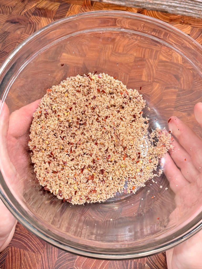A person holds a glass bowl filled with a coarse, reddish-brown crumb mixture, possibly ground nuts or spices, over a wooden surface.