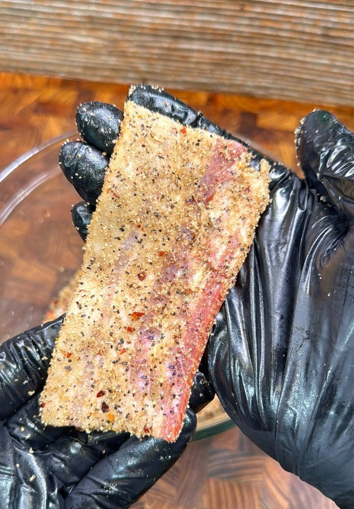 A gloved hand holds a strip of raw bacon coated with a mix of black pepper, crushed red pepper, and seasonings above a glass bowl. A wooden surface is visible in the background.