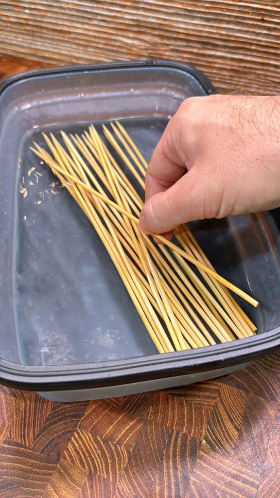 A hand is placing wooden skewers into a shallow plastic container filled with water, likely to soak them before grilling. The container sits on a wooden surface with a striped pattern in the background.