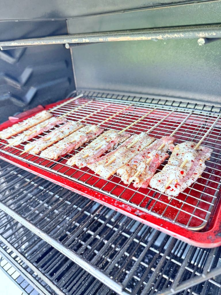 Skewers of seasoned meat are arranged on a red tray inside a barbecue grill with metal racks and dark interior. The meat appears to be marinated and ready to cook.