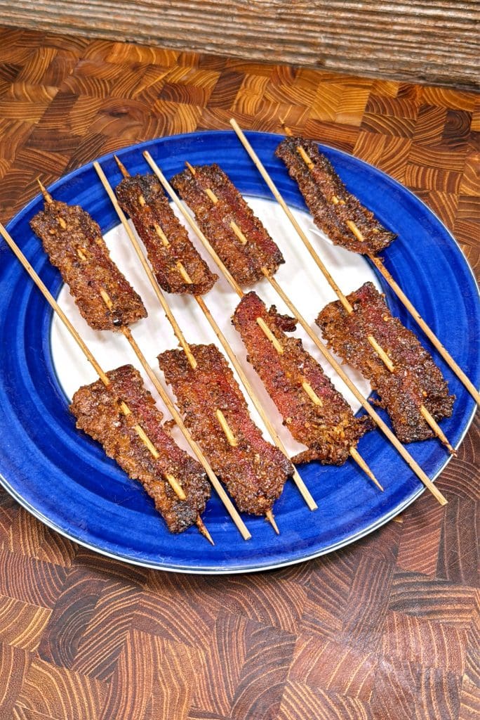 Several skewers of grilled beef suya, a West African spicy meat dish, are arranged on a blue and white plate, which is set on a wooden surface.