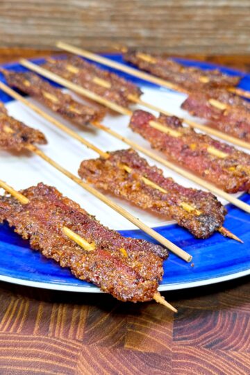 A blue and white plate holds several skewers of grilled, spiced meat arranged neatly on a wooden surface. The meat appears crispy and well-seasoned.