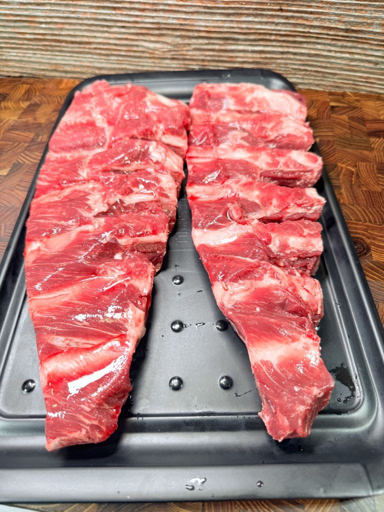 Two racks of raw beef ribs placed side by side on a black tray, showcasing marbled red meat and distinct rib bones, set on a wooden surface with a textured background. Recipe is for beef finger ribs