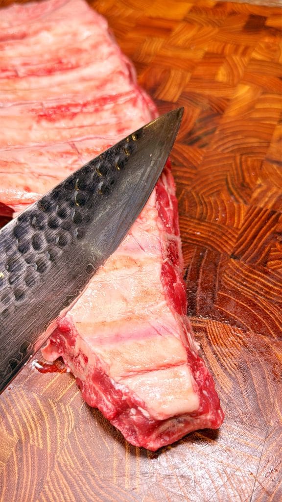 A close-up of a large knife scoring raw beef ribs on a patterned wooden cutting board. The meat and knife are in sharp focus, highlighting the marbling and texture.