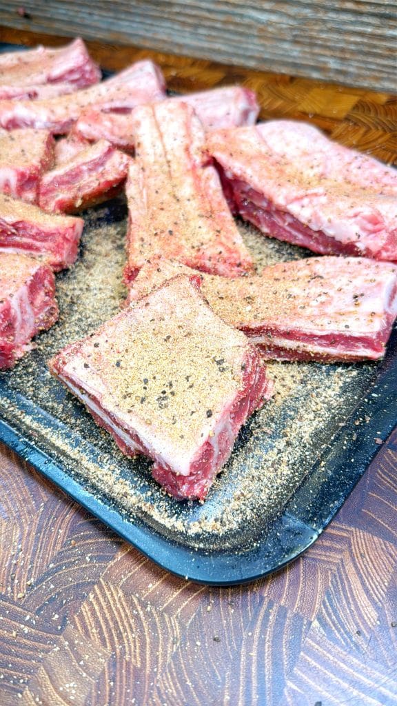 Raw beef ribs coated with seasoning are arranged on a black tray, ready to be cooked. The ribs are covered in a mix of spices and pepper, with some seasoning scattered on the wooden surface.