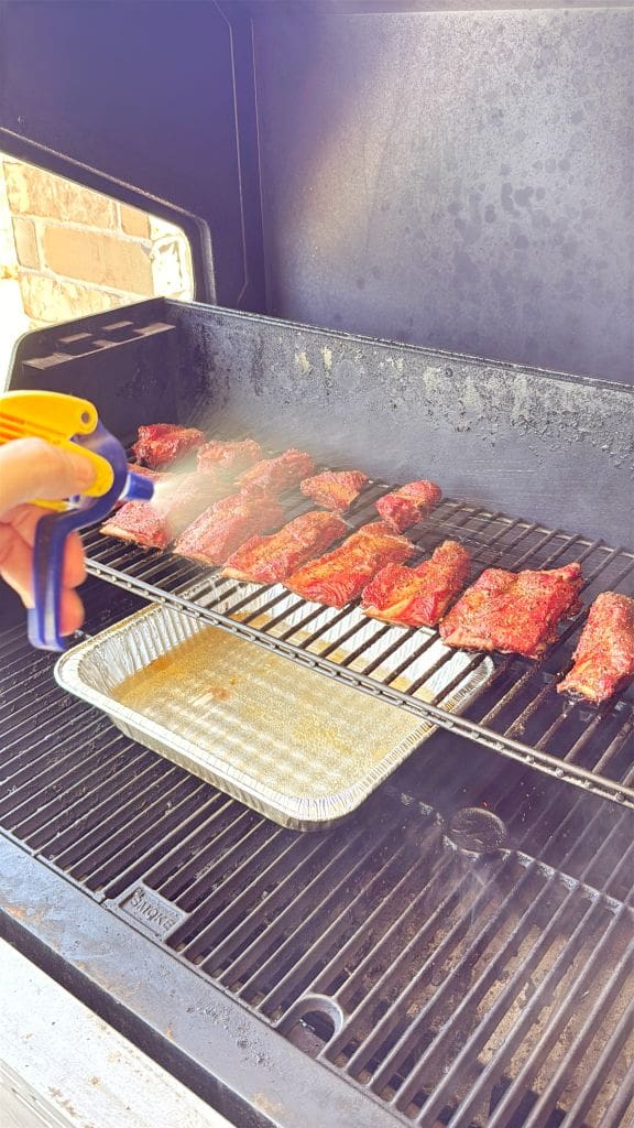 A person sprays liquid onto rows of seasoned meat cooking on a grill, with a disposable tray placed underneath to catch drippings. The scene is outdoors, next to a brick wall.