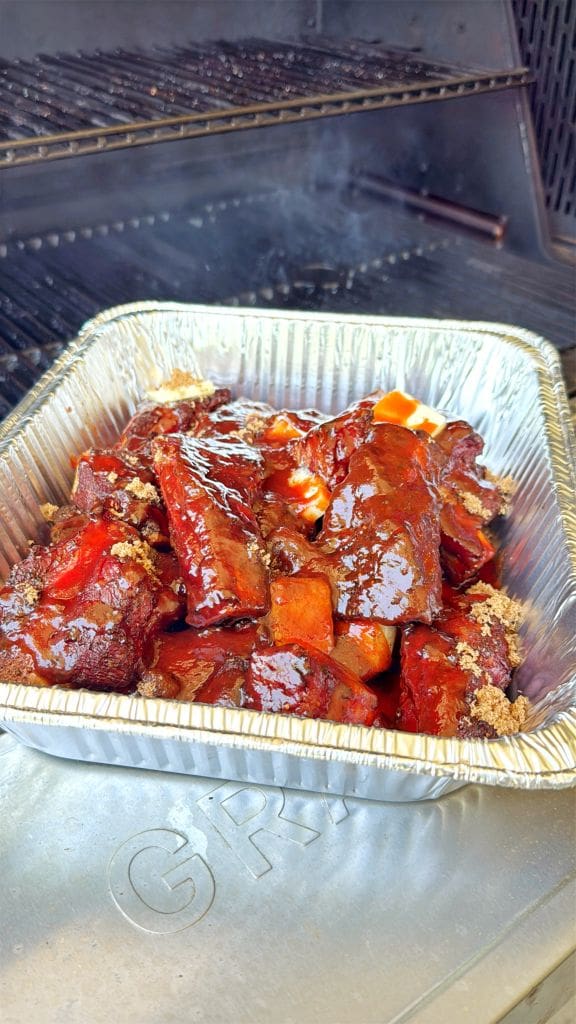 A foil tray filled with glazed, saucy beef ribs sits on the surface of a barbecue grill, with smoke rising from the grill in the background.