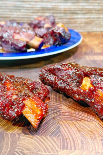 Two glazed, barbecued beef ribs on a wooden cutting board, with a blue plate of more ribs in the background. The ribs are coated in a shiny, dark red sauce.