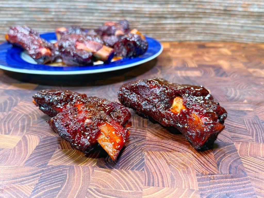 Two saucy, glazed ribs sit on a wooden surface in the foreground, with a blue plate holding more ribs blurred in the background. The ribs have a shiny, caramelized finish.