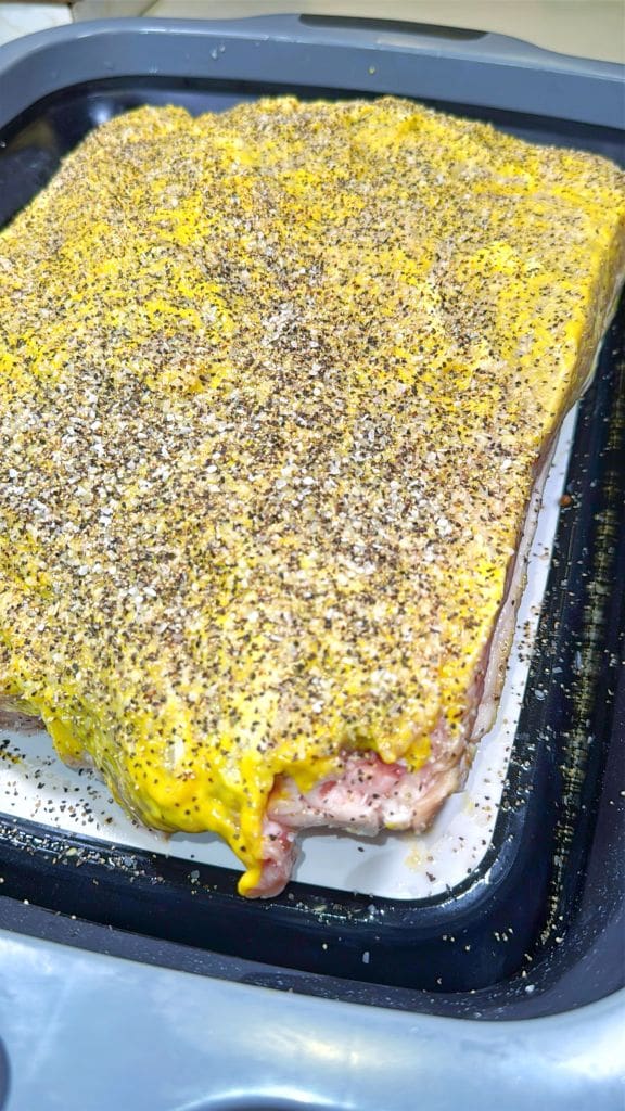 A large rectangular cut of raw meat coated with yellow mustard and heavily sprinkled with cracked black pepper sits on a tray, ready for cooking.