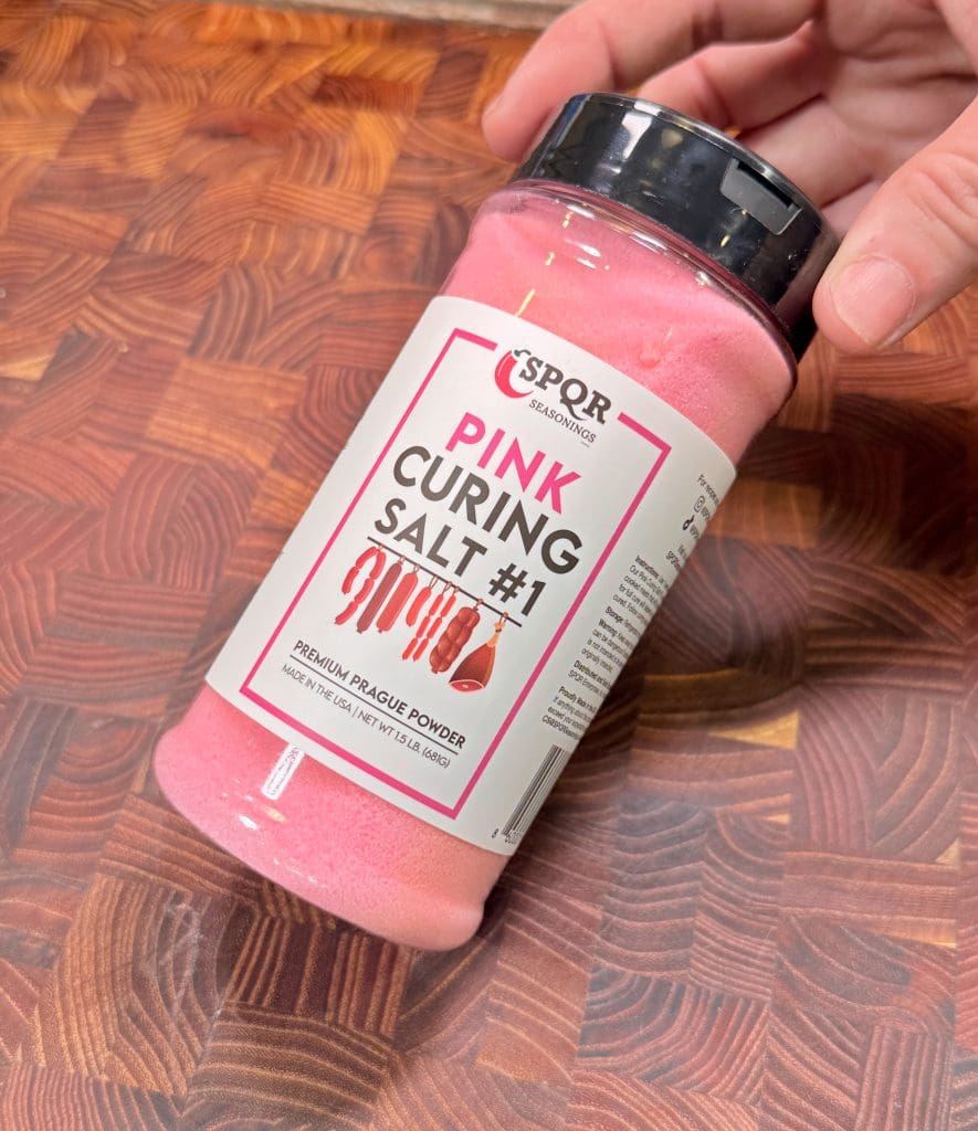 A hand holds a container of SPQR Seasonings Pink Curing Salt #1, labeled Premium Prague Powder, ideal for making pastrami beef ribs, on a wooden surface. The salt inside the container is visibly pink.