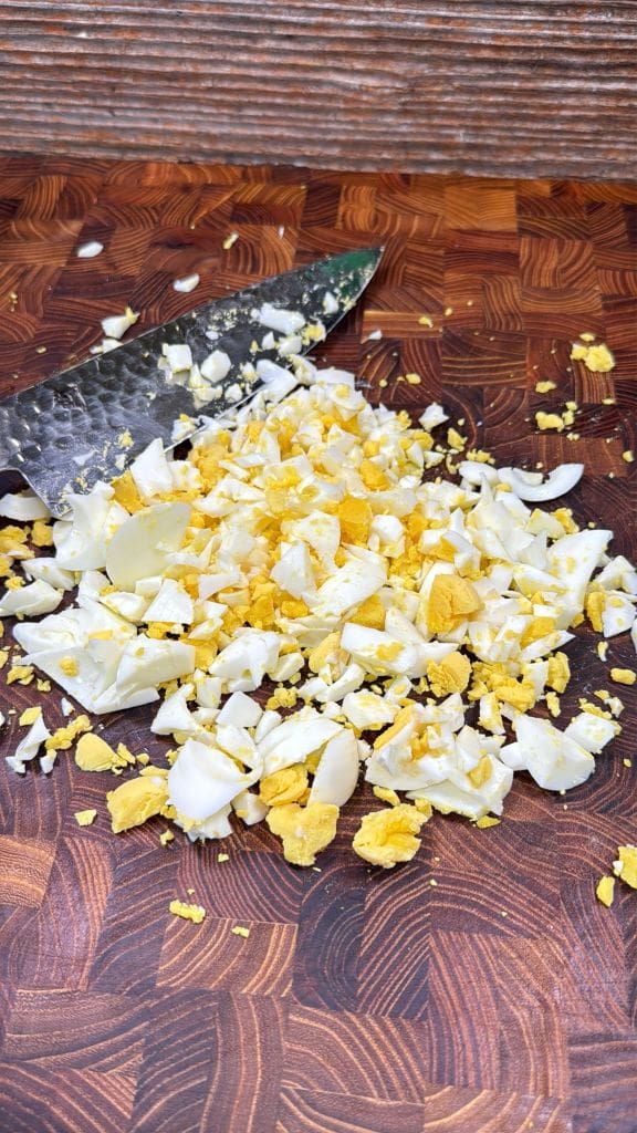 Chopped hard-boiled eggs with yellow yolks and white pieces are spread on a wooden cutting board, with a knife visible beside the eggs.