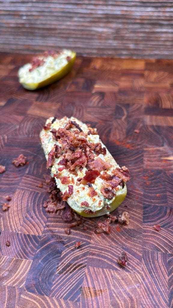 A halved pickle stuffed with creamy filling and topped with crumbled bacon sits on a wooden cutting board, with another stuffed pickle half blurred in the background.