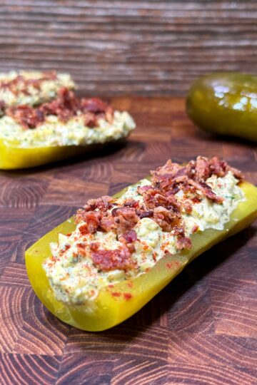 A halved pickle stuffed with a creamy filling and topped with bacon bits, placed on a wooden surface. More stuffed pickles and a whole pickle are in the background.
