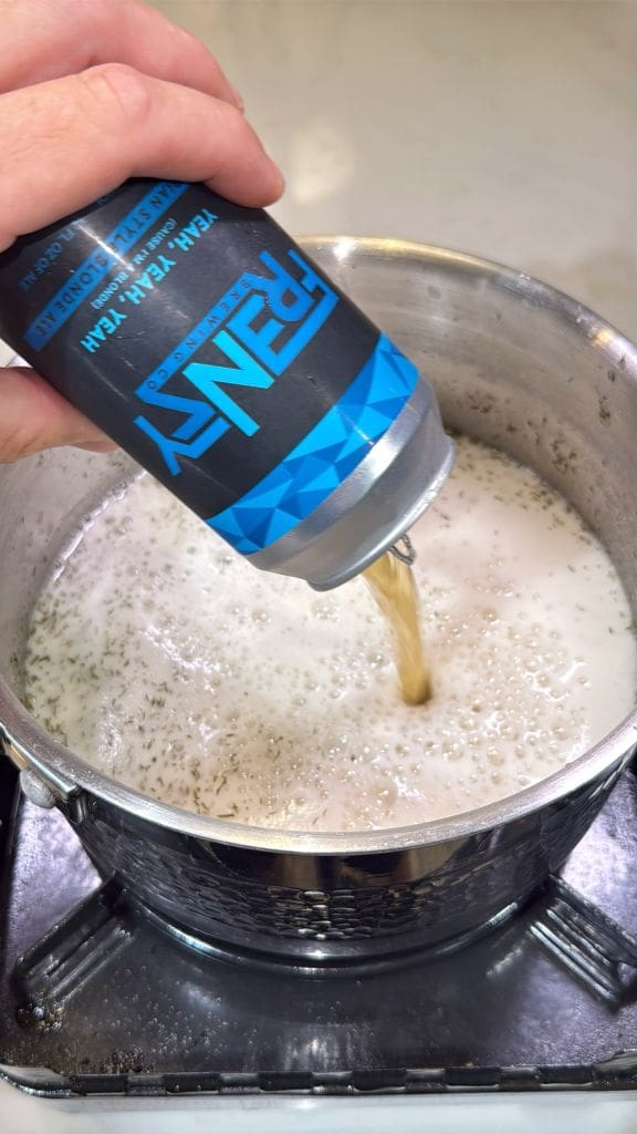 A hand pours a can of Penny IPA beer into a stainless steel pot filled with foamy, bubbling liquid on a stovetop.