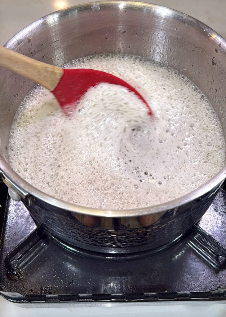 A red spatula stirs a pot of bubbling, frothy liquid on a gas stove, creating a whirlpool effect in the center.