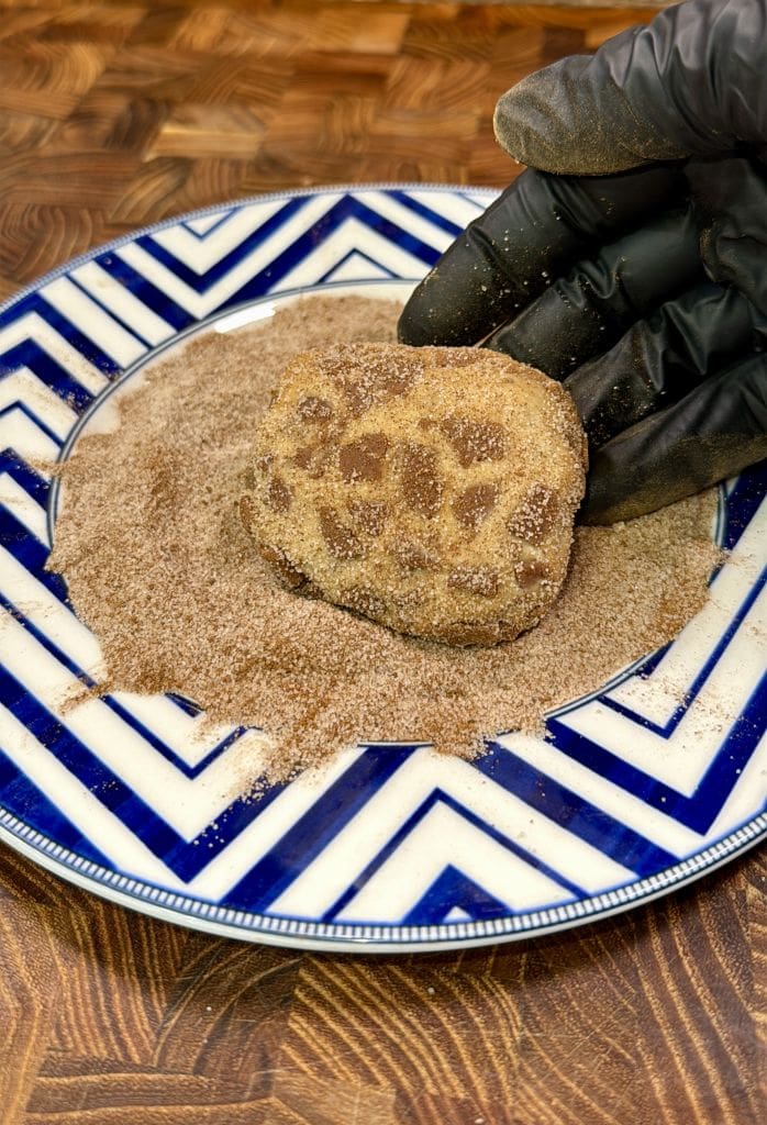 A gloved hand rolls a piece of dough in cinnamon sugar on a blue and white patterned plate, set on a wooden surface.