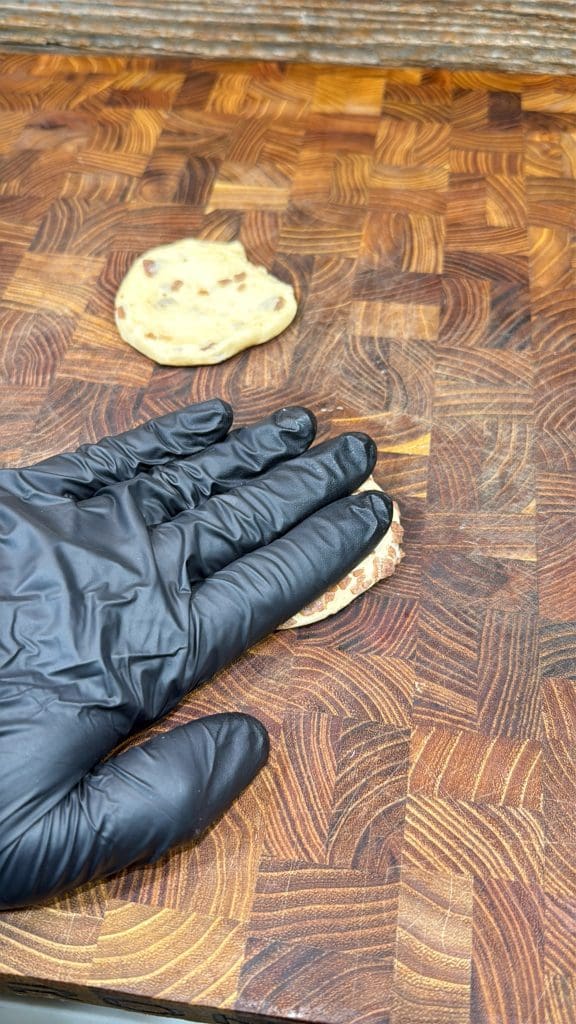 A hand wearing a black glove presses down on a small round piece of dough—soon to become cinnamon roll breakfast bombs—on a wooden cutting board, with another piece of dough in the background.