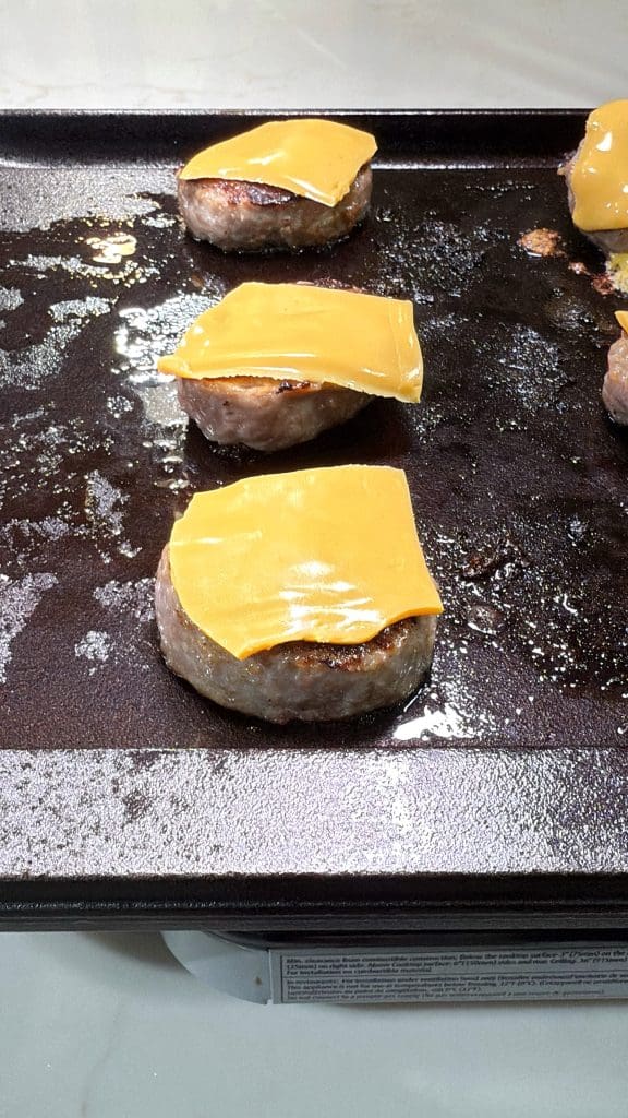Four hamburger patties, each topped with a slice of melted cheddar cheese, are cooking on a greasy griddle. A few more patties and some cinnamon roll breakfast bombs are partly visible on the right side of the image.