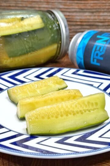 A plate with four pickle spears sits on a wooden surface. Behind the plate, there is a glass jar with pickles and a blue FREN beverage can lying on its side, all against a wood-textured background.