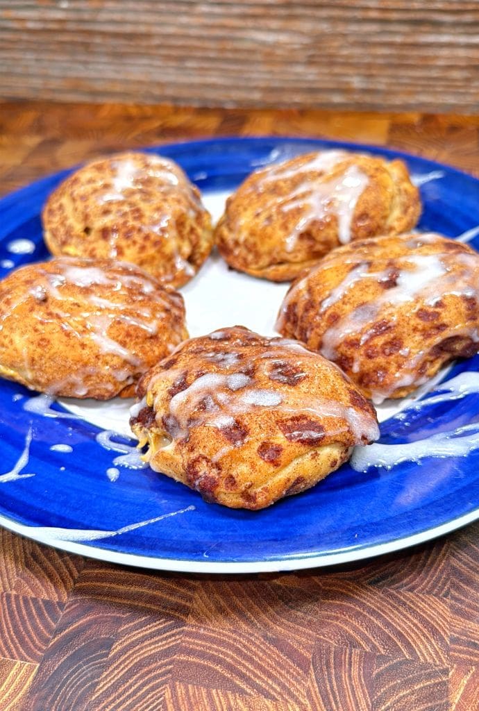Five golden-brown cinnamon rolls with icing drizzle are arranged on a blue and white plate, set on a wooden surface.