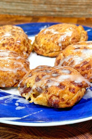 Five iced cinnamon rolls with a drizzle of white glaze are arranged on a blue and white plate, set on a wooden surface with a wood-patterned background.