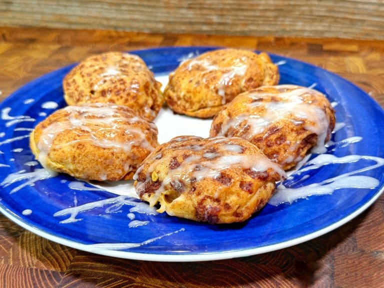 Five iced cinnamon rolls with a drizzle of white glaze are arranged on a blue and white plate, set on a wooden surface with a wood-patterned background.