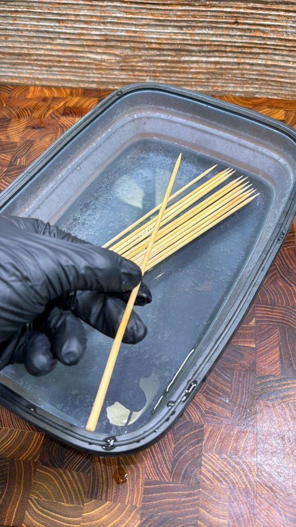 A gloved hand holds a wooden skewer above a plastic container filled with water, where several skewers are soaking on a wooden surface.