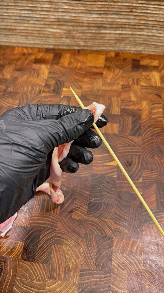A hand wearing a black glove holds a piece of raw bacon, wrapping it around a wooden skewer on a patterned wooden surface.