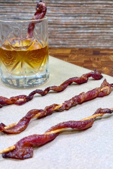 A glass of whiskey with a slice of bacon inside sits on a napkin, next to three skewers of twisted, cooked bacon on a wooden surface.