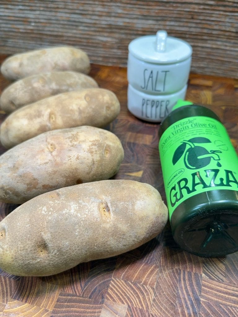 Five russet potatoes on a wooden surface with a container of Graza extra virgin olive oil and a small ceramic jar labeled Salt Pepper in the background.